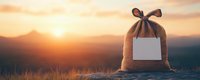 Canvas gift bag with blank label against scenic mountain sunset panorama, warm golden light illuminating rustic burlap texture in nature setting.
