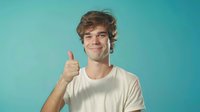 Young Caucasian man with wavy brown hair wearing white t-shirt showing thumbs up gesture against turquoise background, expressing positive attitude and approval.