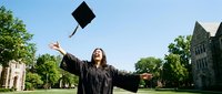 Young woman in graduation gown celebrating achievement by throwing mortarboard cap in air on university campus with historic buildings and trees.