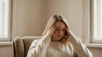 Young blonde woman experiencing headache or stress sitting on beige armchair by window in cozy living room interior with natural lighting.