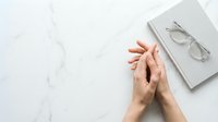 Female hands clasped together in prayer position on white marble surface with notebook and eyeglasses, symbolizing mindfulness and reflection.
