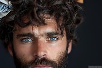 Close up portrait of young bearded man with intense blue eyes and dark curly hair against black background, dramatic lighting emphasizes facial features.