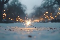 Burning sparkler in snow creates magical winter atmosphere with blurred bokeh lights and trees in background, festive holiday celebration moment captured at dusk.
