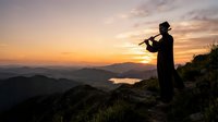 Silhouetted musician playing flute on mountain peak at sunset overlooking valley and lake below, suitable for music, nature, and inspirational content.