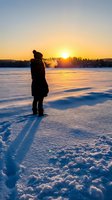 Woman standing on frozen lake watching golden sunrise over snowy landscape. Winter solitude and nature contemplation scene for travel, wellness, and seasonal content.