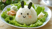 Cute bunny-shaped rice ball with nori seaweed ears and facial features served on fresh lettuce leaves, creative Asian food styling for children's meal or bento lunch.