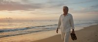 Senior man with gray hair and light skin tone, wearing casual beige linen clothing, walking barefoot on sandy beach at sunset holding shoes, contemplative lifestyle portrait.