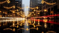 Urban street at night with golden holiday lights reflecting on wet pavement, creating magical mirror effect with light trails from passing traffic in city center.