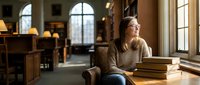 Young woman student relaxing in university library with books, studying in academic environment with natural window light and warm atmosphere.