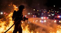 Firefighter silhouette battling intense flames at night with city lights in background. Emergency response scene with dramatic fire and smoke effects.