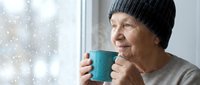 Senior woman in knit hat enjoying hot beverage by snowy window, cozy winter morning scene for lifestyle and wellness content.