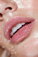 Close-up of glossy pink lips with water droplets on smooth skin, showing white teeth. Macro beauty shot highlighting moisturized lips and fresh complexion.
