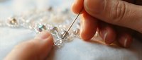 Hands threading sparkling crystal beads with a needle on white fabric, creating elegant handmade jewelry in a close-up craft scene.