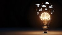 Glowing light bulb with brain inside and business icons floating above on dark background representing innovation and creative thinking concepts.