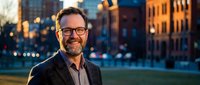 Professional middle-aged Caucasian businessman with glasses and beard smiling confidently in urban downtown setting during golden hour for corporate headshots.