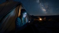 Young man with light skin tone camping under starry night sky, holding glowing tablet device inside tent with milky way galaxy visible above landscape at dusk.