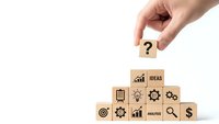 Hand placing wooden cube with question mark on pyramid of business icon blocks representing strategic planning and decision making process.