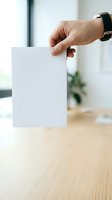 Male hand holding blank white card in modern office environment with wooden desk and blurred background, suitable for business branding and marketing mockups.