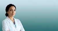 Young woman with dark hair and South Asian ethnicity wearing white lab coat, arms crossed, smiling confidently against teal gradient background. Healthcare professional portrait.