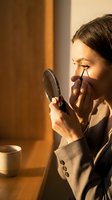 Young Caucasian businesswoman applying mascara with a hand mirror in warm morning light, seated at a wooden desk with a coffee cup.