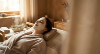 Young woman relaxing with facial mask in cozy bedroom during morning skincare routine. Self-care wellness concept for spa and beauty treatments.