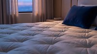 Cozy bedroom with quilted white bedding, navy blue pillow, and ocean view at twilight through window with curtains.