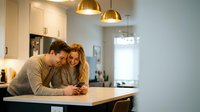 Happy young Caucasian couple using smartphone together in modern kitchen with white cabinets and gold pendant lights for lifestyle content.