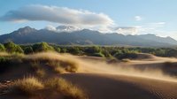 Desert sand dunes with golden grass tufts at sunrise, dramatic mountain range backdrop under cloudy sky, natural wilderness landscape in warm morning light.
