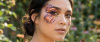 Young woman with artistic monarch butterfly face painting covering left eye area in natural outdoor garden setting for creative makeup and body art concepts.