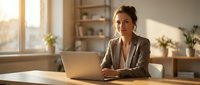 Professional businesswoman working on laptop in modern home office with natural sunlight streaming through window creating warm productive workspace atmosphere.