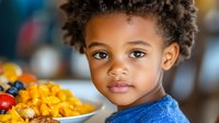 Young African American toddler boy with curly hair looking at camera while eating colorful breakfast cereal in kitchen setting for family lifestyle content.