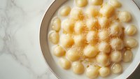 Homemade potato gnocchi dumplings arranged in circular pattern on white ceramic plate with marble background, sprinkled with nutmeg and parmesan cheese.