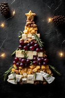 Christmas tree shaped cheese board with assorted cheeses, grapes, nuts, and rosemary on dark slate background with pine cones and string lights.
