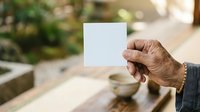 Senior man with brown skin holding blank white card outdoors in garden setting, suitable for greeting cards, invitations, and promotional messaging.
