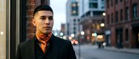 Young Hispanic businessman in elegant black coat and orange scarf standing confidently on urban city street at dusk with blurred lights and brick buildings.