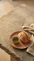 Freshly baked bread roll with golden crust served on terracotta plate alongside small bowl of olive oil, styled on rustic stone surface with neutral linen fabric for food photography and culinary content.