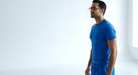 Man in blue athletic shirt and shorts wearing glasses, standing in profile against white background, suitable for fitness, sportswear, and wellness marketing.