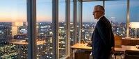 Mature businessman in formal suit standing at floor-to-ceiling windows overlooking illuminated cityscape at dusk, contemplating urban skyline from high-rise office building.