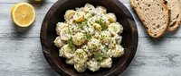 Creamy potato salad with fresh herbs and lemon served in dark bowl with bread slices on rustic wooden table for lunch or dinner recipes.