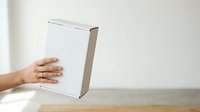 Caucasian male hand holding white cardboard box on wooden surface against neutral background for product packaging mockup and shipping display.