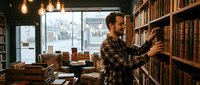 Young man with dark hair and beard, wearing plaid shirt, selecting book from wooden shelf in cozy library with pendant lights, leather armchair, and city skyline view through window.