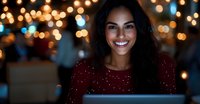 Young Latin woman with bright smile working on laptop at night, illuminated by warm bokeh lights and screen glow in festive atmosphere.