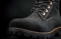 Close-up view of black leather work boot with thick rubber sole, contrast stitching and metal eyelets against dark background, showcasing rugged construction.