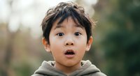 Young Asian boy with surprised expression wearing gray hoodie outdoors with natural lighting and soft background blur for lifestyle photography.