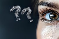 Close-up of brown female eye with long eyelashes next to hand drawn question marks on dark background, representing curiosity, doubt and questioning.