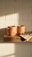 Two handmade ceramic mugs on rustic wooden shelf against white brick wall with warm natural lighting and eucalyptus branch for cozy home decor.
