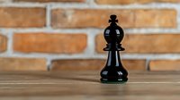 Black bishop chess piece on wooden table against brick wall background, selective focus creates dramatic composition for business strategy concepts.