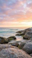 Serene coastal landscape at sunrise with moss-covered boulders lining sandy beach, turquoise waves, and soft pastel sky in pink and gold tones. Ideal for travel and nature projects.