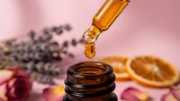 Essential oil dropper bottle with golden liquid drop, lavender flowers and dried orange slice on pink background for aromatherapy and natural wellness.