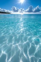 Sunlight sparkles on crystal clear turquoise ocean water with white sandy beach and fluffy clouds on horizon, creating mesmerizing light patterns on surface.